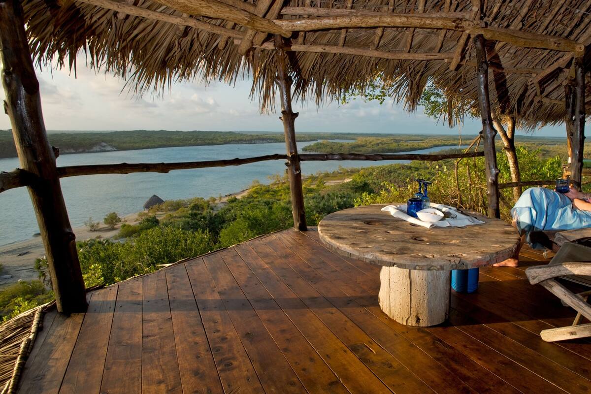 Una terraza de madera con techo de paja que da a un cuerpo de agua y vegetación.