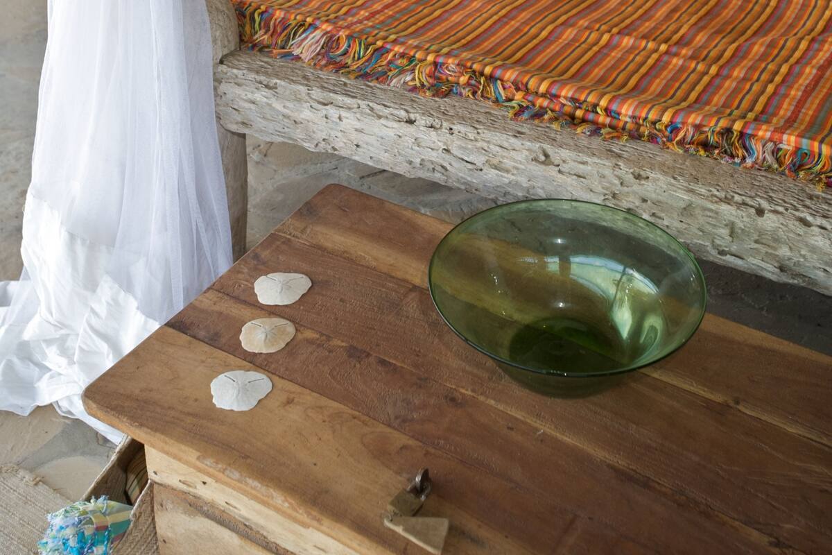 Una cama con una manta a rayas, un cuenco verde y una mesa de madera con conchas.
