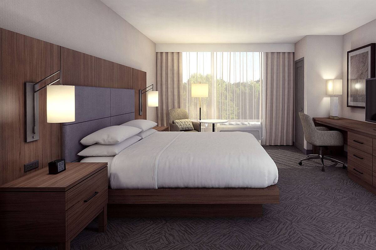 A hotel room with a large bed, a desk with a TV, a chair, a small table, and a window with curtains.