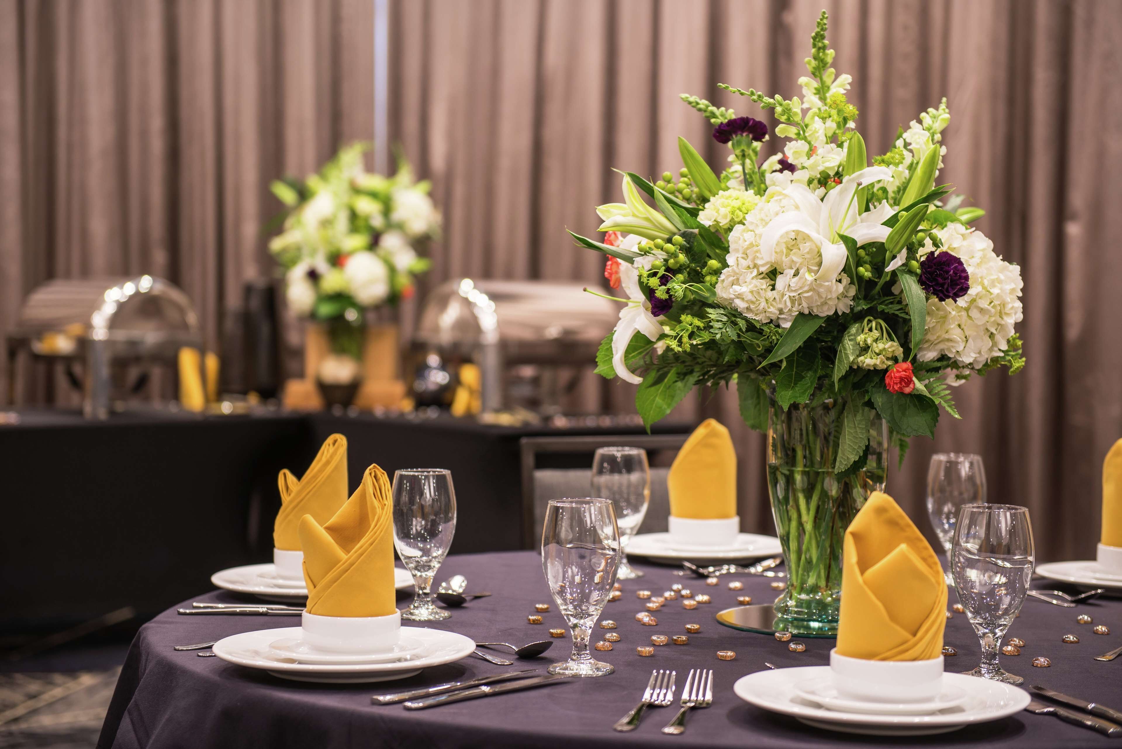 A dining table set with white plates, gold napkins, and clear glassware, surrounded by a floral centerpiece.