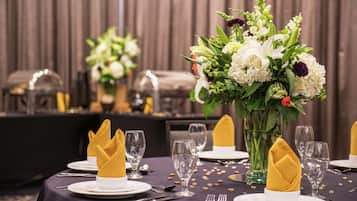 A dining table set with white plates, gold napkins, and clear glassware, surrounded by a floral centerpiece.