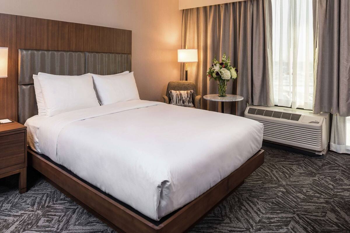 A hotel room with a large bed, a nightstand, a small table with a vase of flowers, a wall-mounted heater, and a window with curtains.