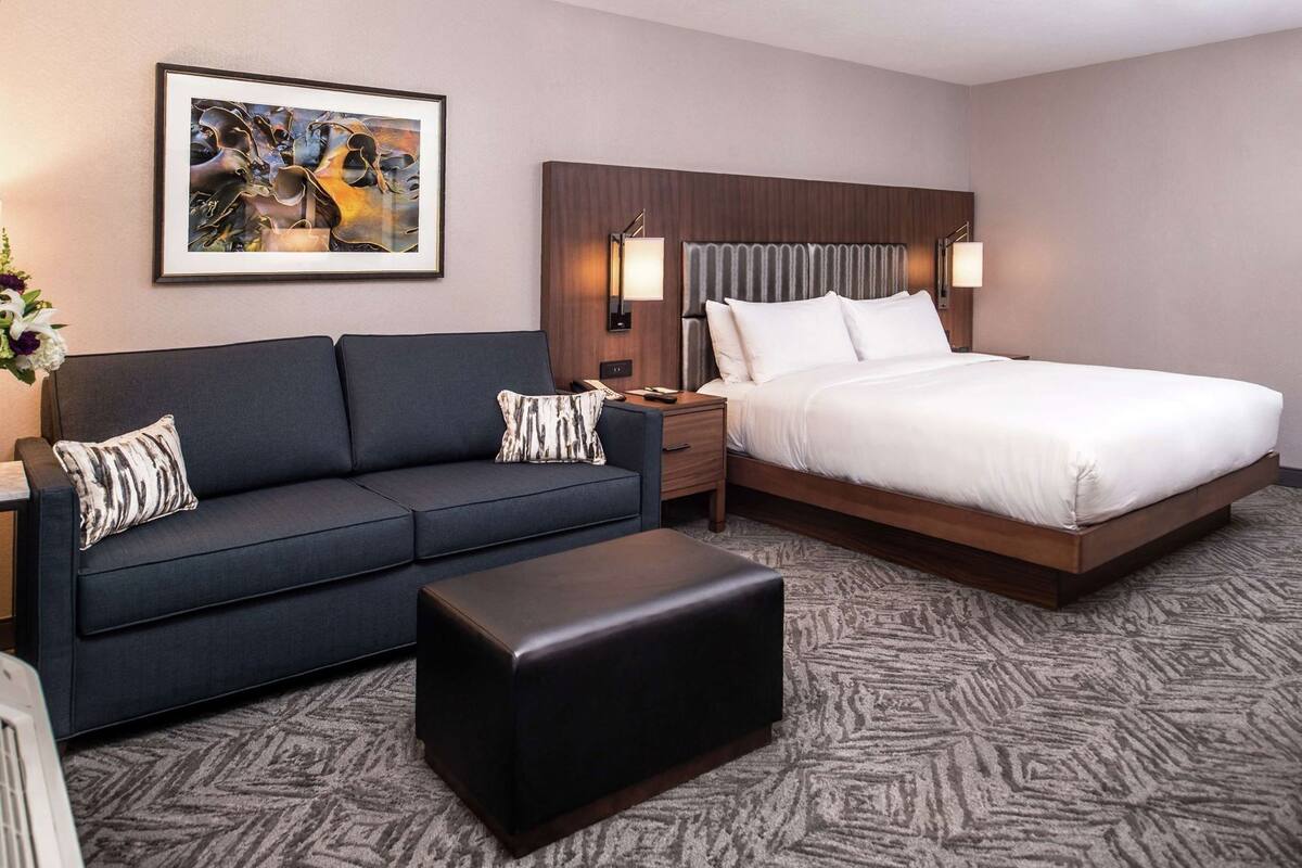 A hotel room with a bed, a sofa, an ottoman, a nightstand, a lamp, and a framed picture on the wall.