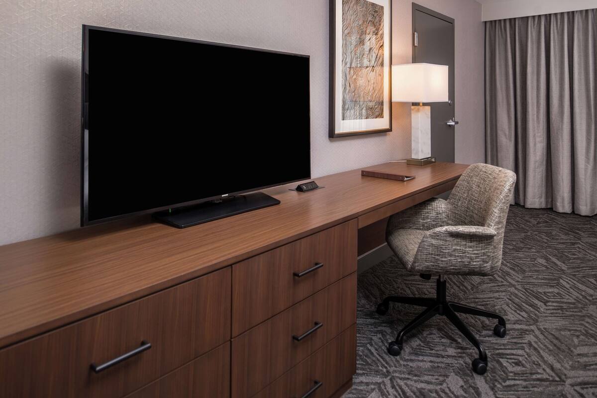 A hotel room with a large flat-screen TV on a wooden desk, a chair, a lamp, and a framed artwork on the wall.