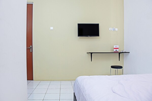 Una habitación de hotel con una cama, un televisor montado en la pared, un taburete pequeño y una estantería con una caja.