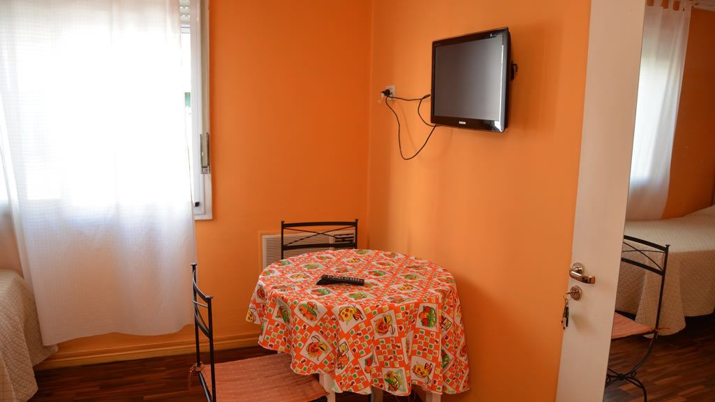 A room with an orange wall, a round table with a patterned tablecloth, a television mounted on the wall, and a bed with a white bedspread.