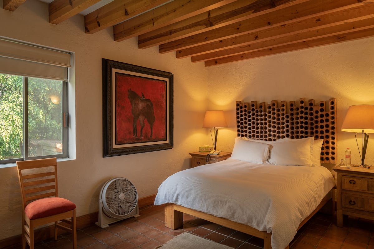 Une chambre avec un lit en bois, deux lampes de chevet, un tableau encadré au mur, une chaise et un ventilateur sur pied.