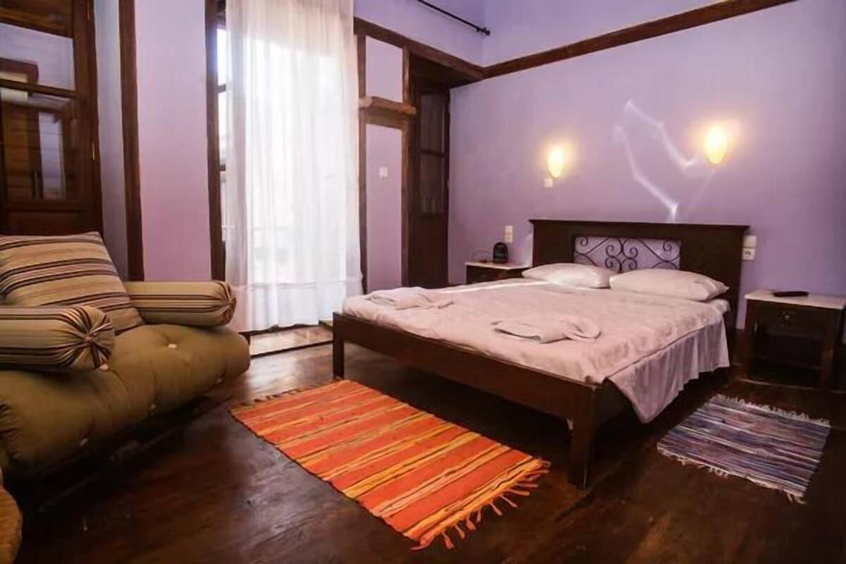 Un dormitori amb un llit, una cadira, una nitzeta i una finestra amb cortines.