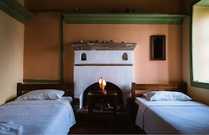 A room with two beds, a fireplace, and a small table with a lamp. - Karnayo Traditional Houses (Kastellorizo)