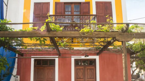 A two-story building with a yellow facade, a balcony with a wooden railing, and a stone entrance. - Karnayo Traditional Houses (Kastellorizo)