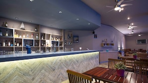 Een bar met flessen en glazen op planken, een marmeren toonbank, houten tafels en plafondventilatoren.