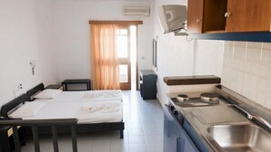 Ein kleines Hotelzimmer mit einem Einzelbett, einem Nachttisch, einer Küchenzeile mit Herd und Spüle sowie einem Fenster mit Vorhängen.