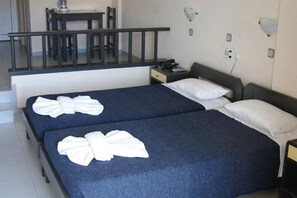 A hotel room with two beds, each with a blue bedspread and white pillows. There is a small bedside table with a lamp and a phone. A desk and chair are visible in the background. - Relax Hotel (Chania)
