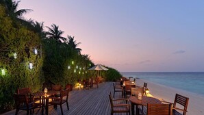 An outdoor dining area with wooden tables and chairs, string lights, and a view of the ocean at dusk. - Embudu Village (Emboodhoo)