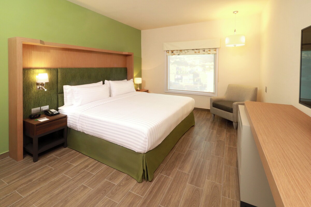 A hotel room with a bed, bedside table, lamp, chair, and a window with a view.