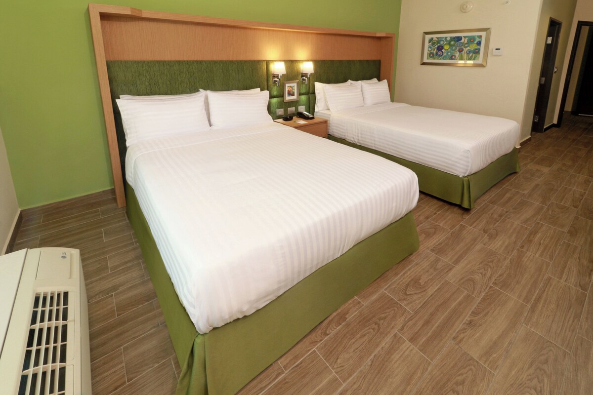 A hotel room with two beds, a nightstand with a phone and lamp, a wall-mounted picture, and a visible air conditioning unit.