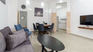 A modern living room with a grey sofa, a dining table with chairs, a TV mounted on the wall, and a kitchen area in the background. - Liber Seashore Suites Aparthotel (Tel Aviv)