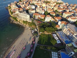 Vista aérea de un pueblo costero con una playa, un edificio grande de fachada blanca y una zona densamente poblada de viviendas.