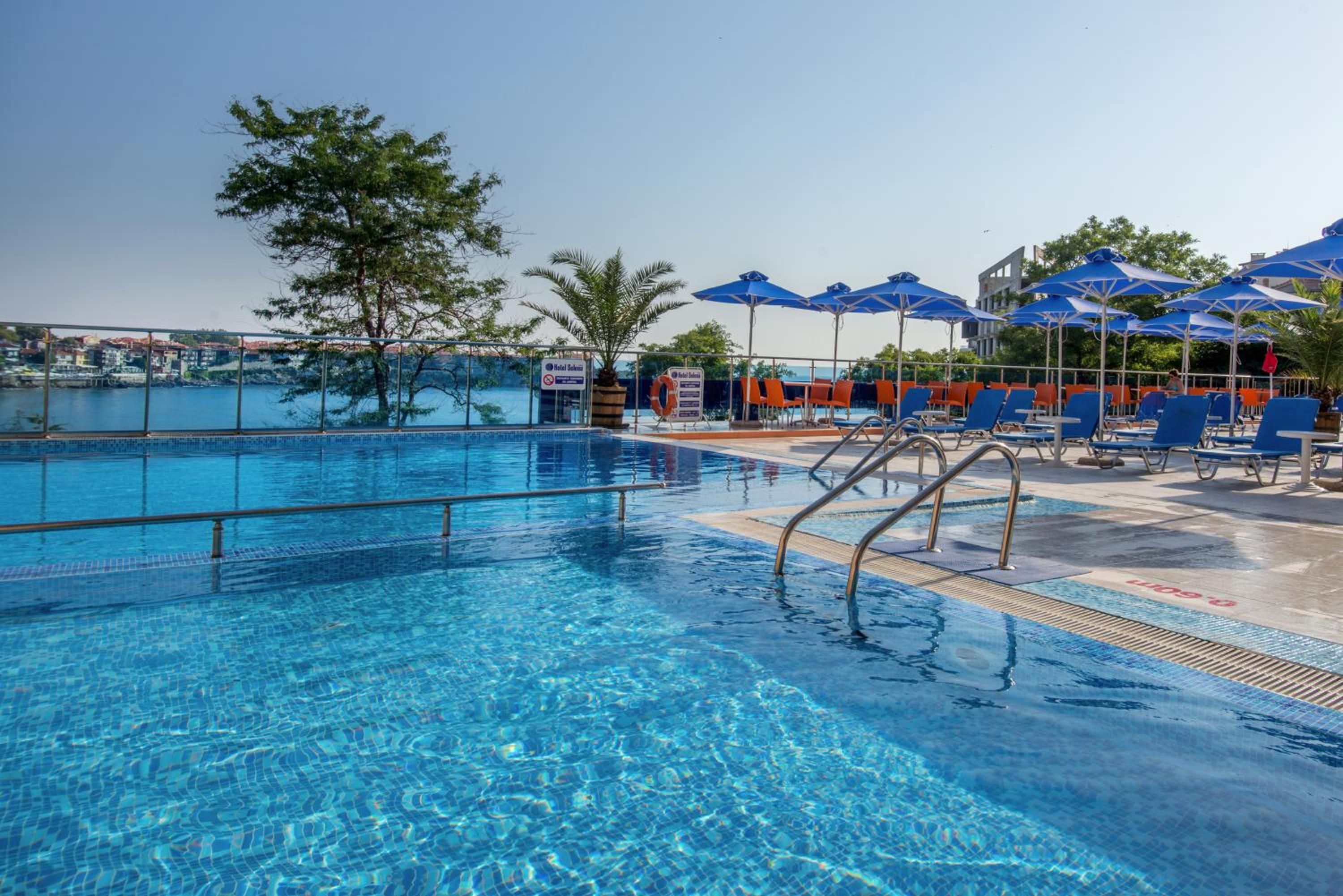 Ein klares Schwimmbecken mit blauem Wasser, umgeben von Liegestühlen und Sonnenschirmen, mit Blick auf eine Küstenstadt im Hintergrund.