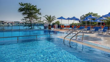 Ein klares Schwimmbecken mit blauem Wasser, umgeben von Liegestühlen und Sonnenschirmen, mit Blick auf eine Küstenstadt im Hintergrund.