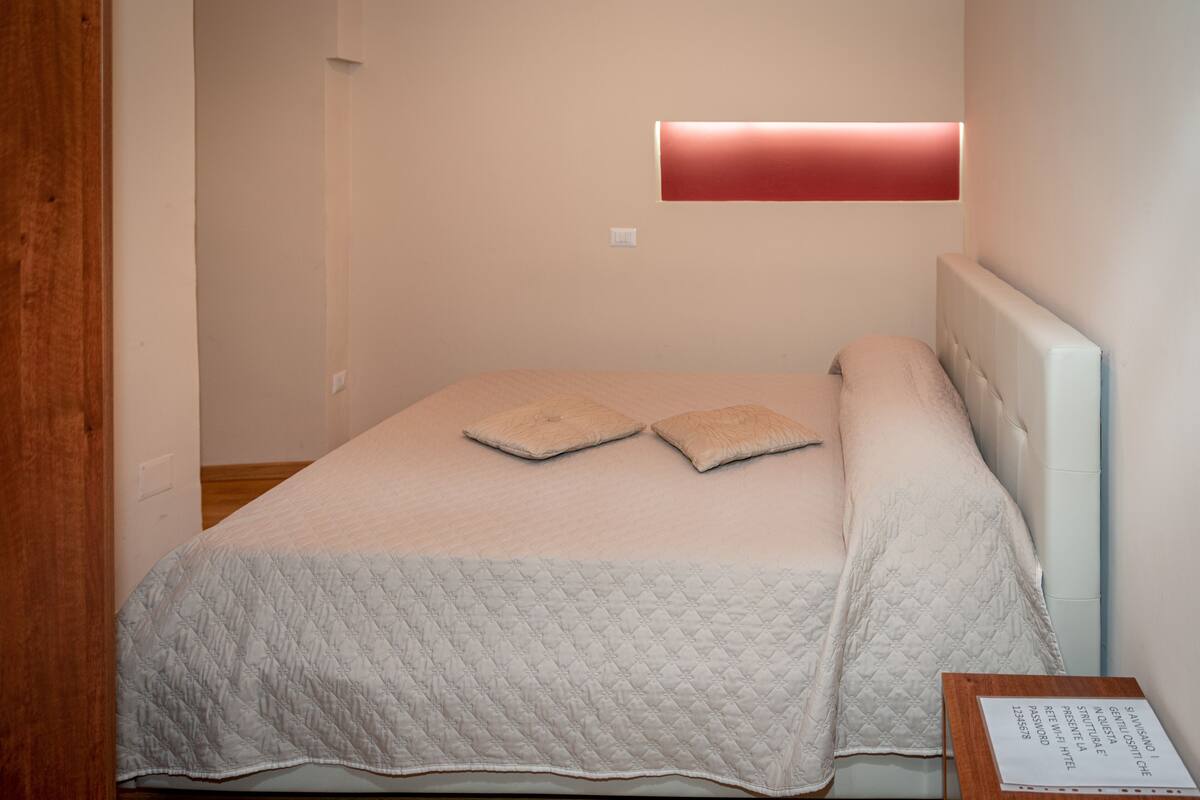 Un lit bien fait, avec une couvre-lit matelassé et deux oreillers, une table de chevet sur laquelle est posé un petit panneau, et un luminaire fixé au mur.