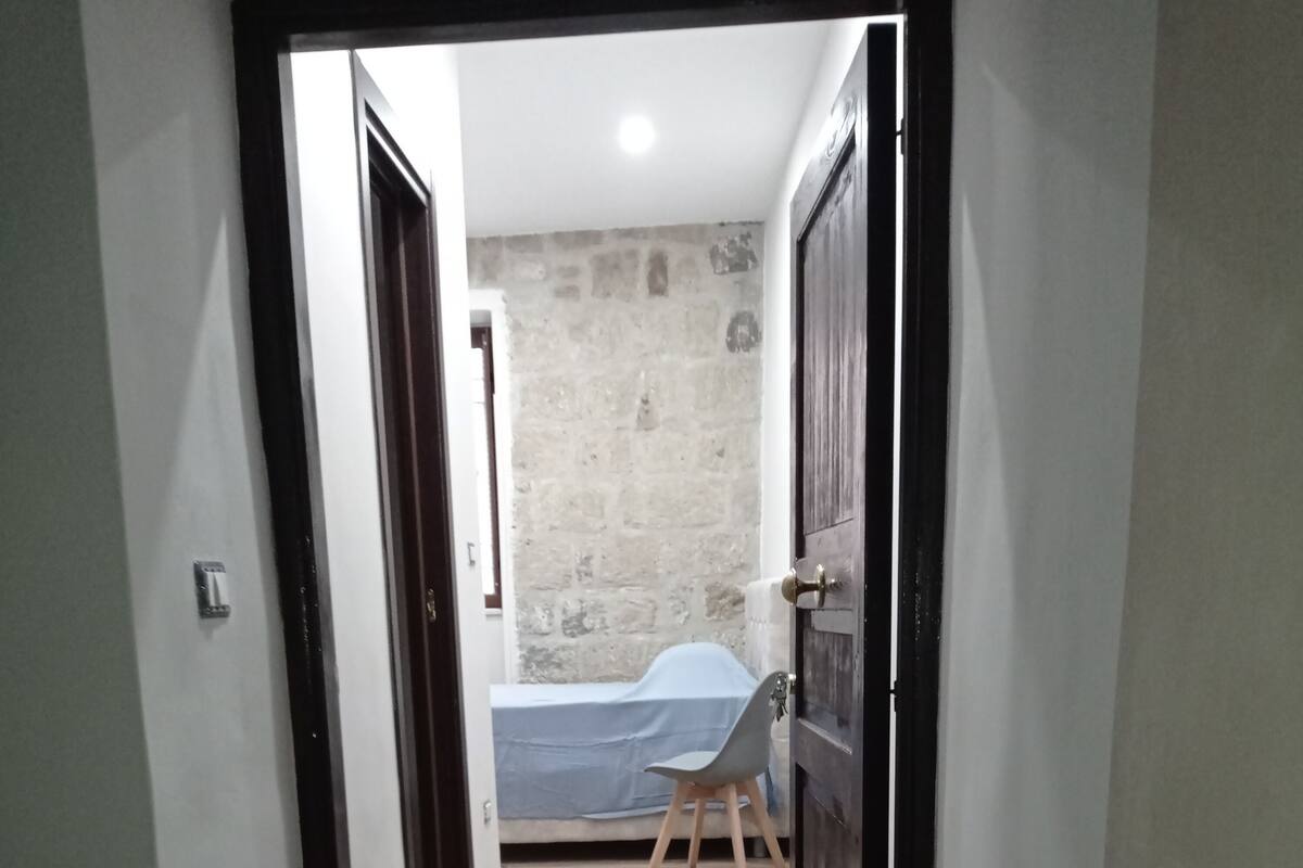 Un couloir étroit mène à une pièce où se trouvent un lit, une chaise et un mur en pierre.