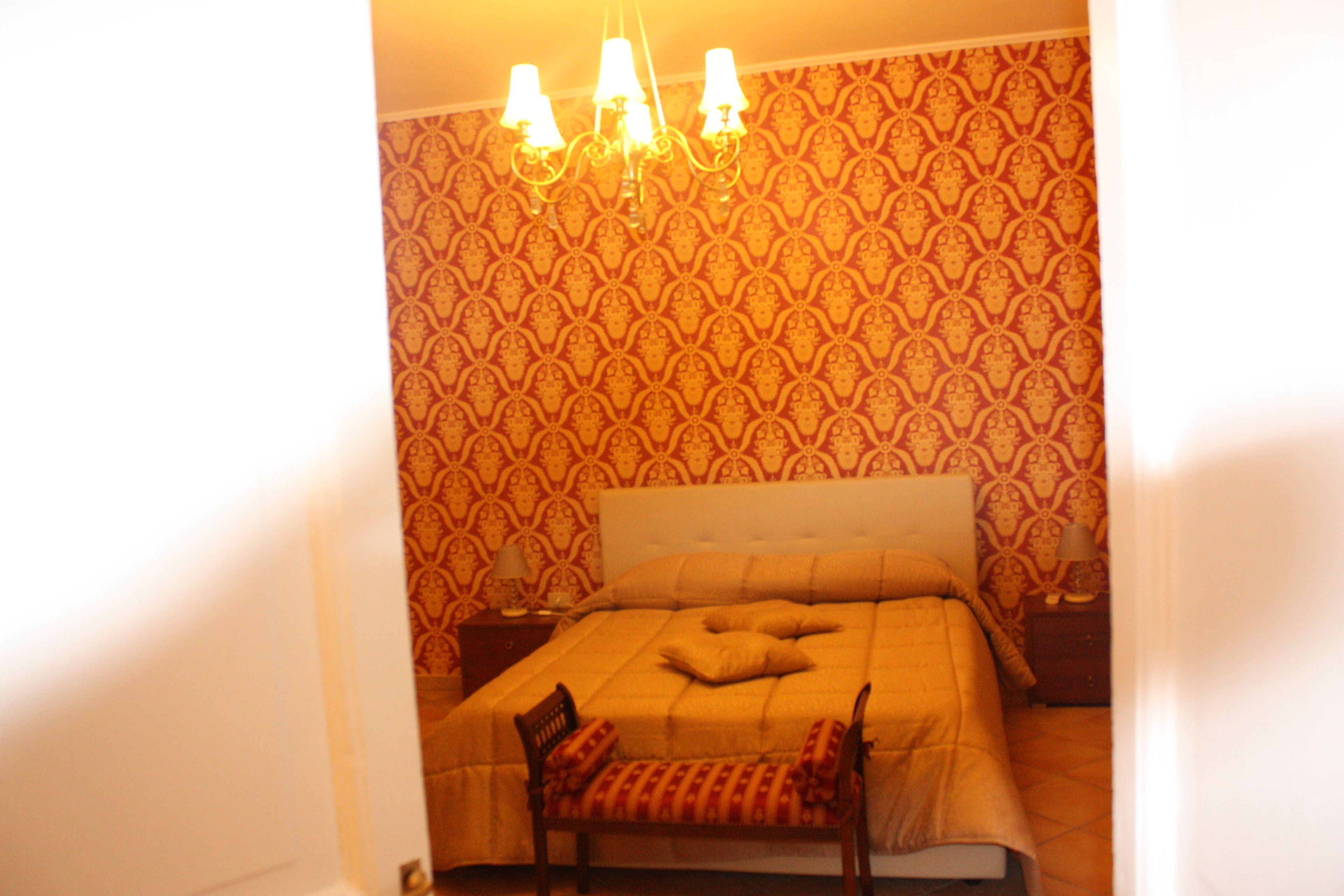 Une chambre avec un papier peint à motifs, un lit recouvert d’une couette beige, une table de chevet avec une lampe et une chaise agrémentée d’un coussin à motifs.