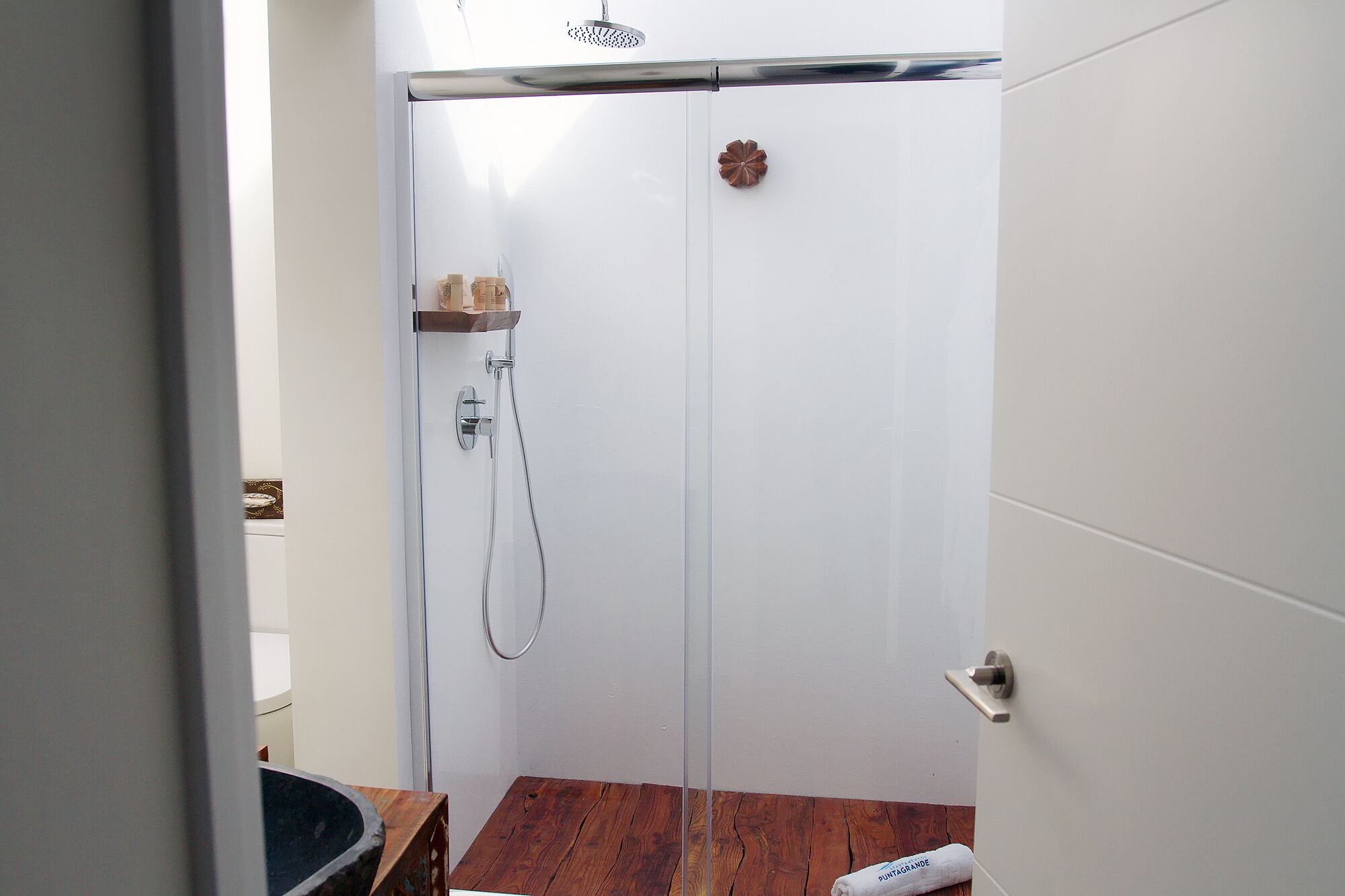 Een witte badkamer met een douche, een houten plank met toiletartikelen en een handdoek op de vloer.
