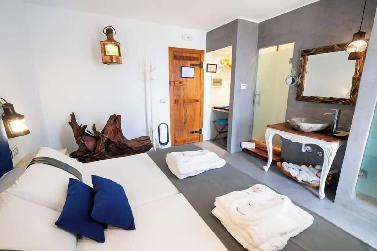 A modern bedroom with a bed, bedside lamps, a wooden sculpture, a bathroom with a sink and mirror, and a door leading to another room.