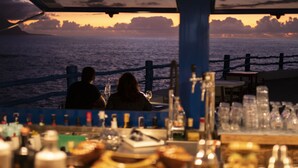 Un bar con vistas al océano al atardecer, que incluye una barandilla y varias botellas y vasos.