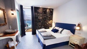 Un dormitorio moderno con una pared de acento de piedra, una cama grande con ropa de cama azul, una mesita de noche y vistas al mar.