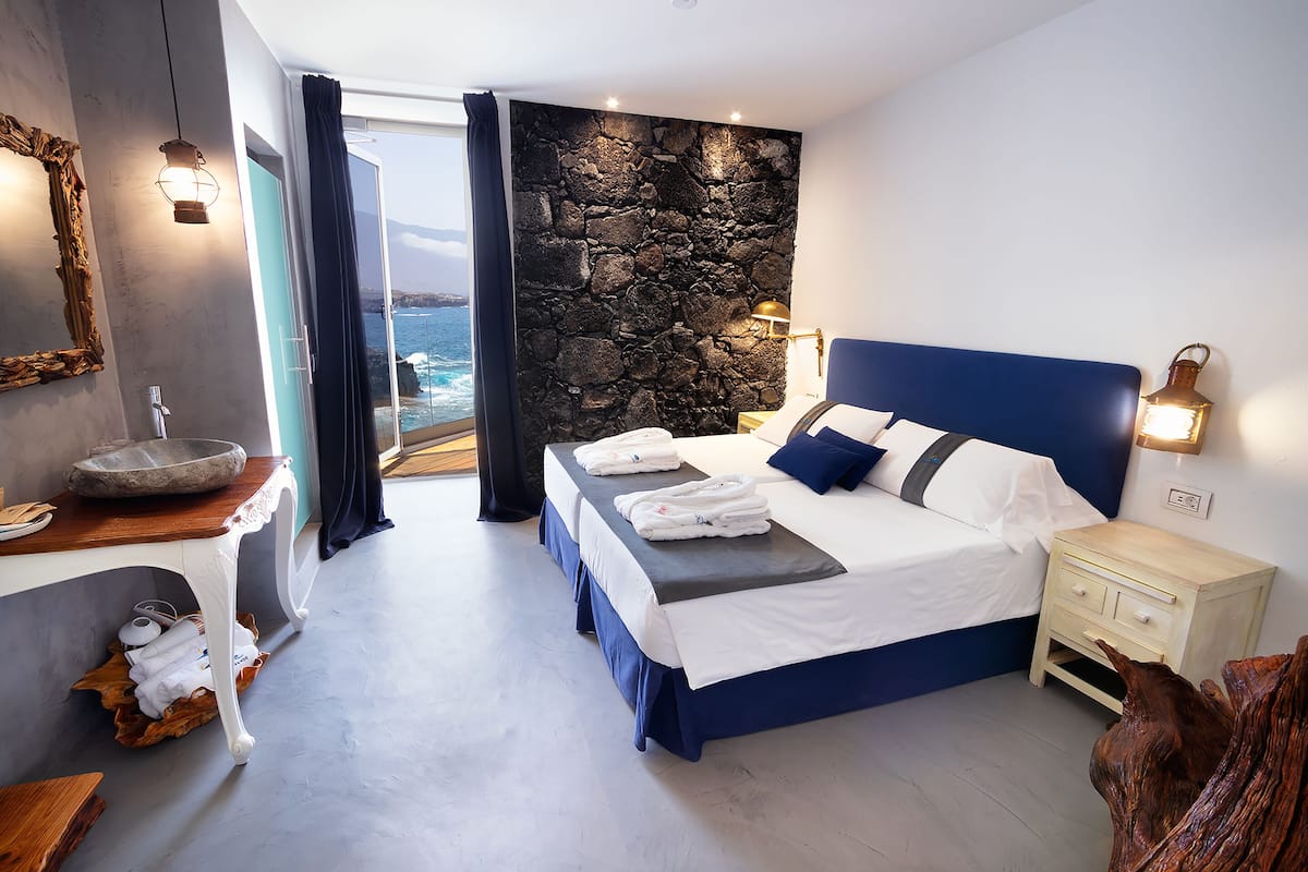 A modern bedroom with a stone accent wall, a large bed with blue bedding, a nightstand, and a view of the sea.