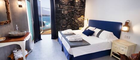 A modern bedroom with a stone accent wall, a large bed with blue bedding, a nightstand, and a view of the sea.