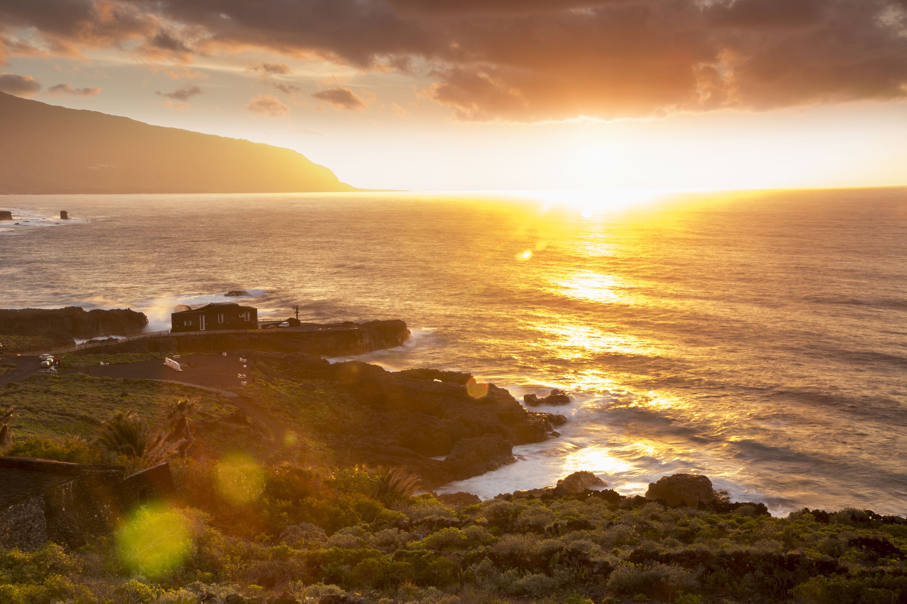 A coastal landscape with a sunset over the ocean, rocky terrain, and a small building near the shore.