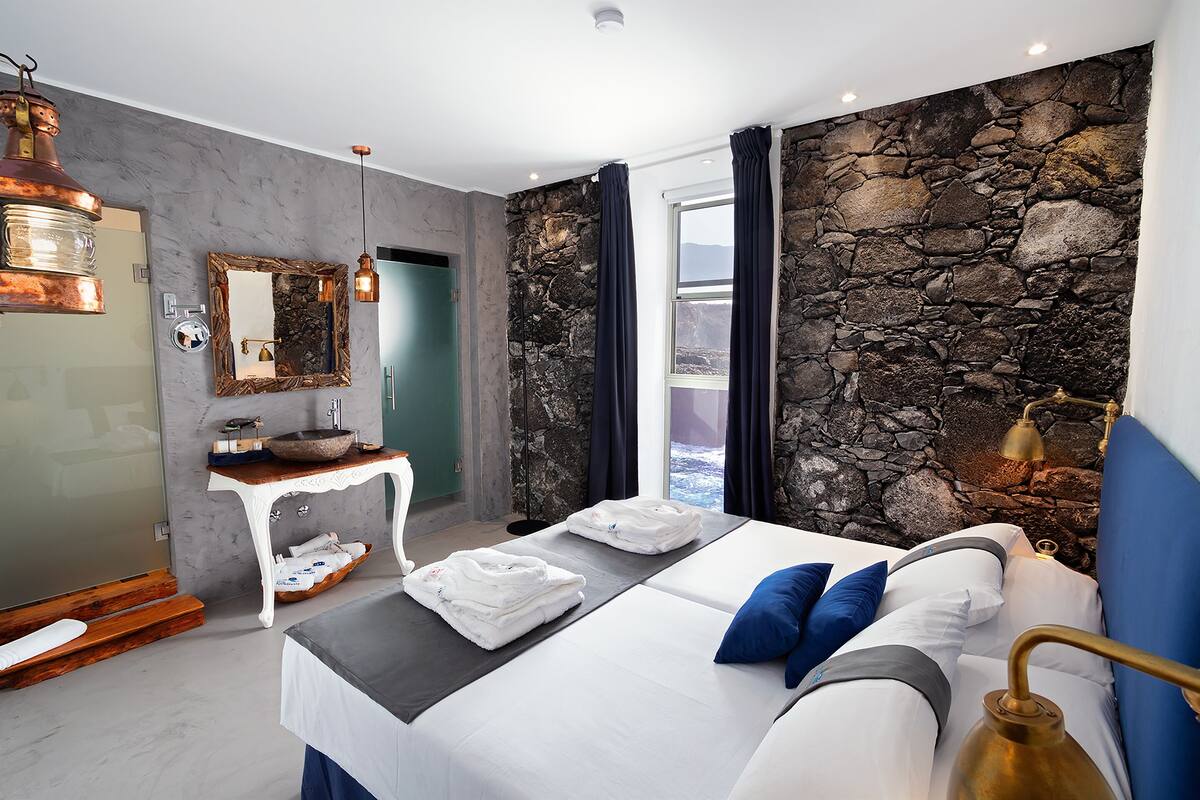 A modern hotel room with a large bed, a stone accent wall, a small table with a mirror, and a view of the outdoors.
