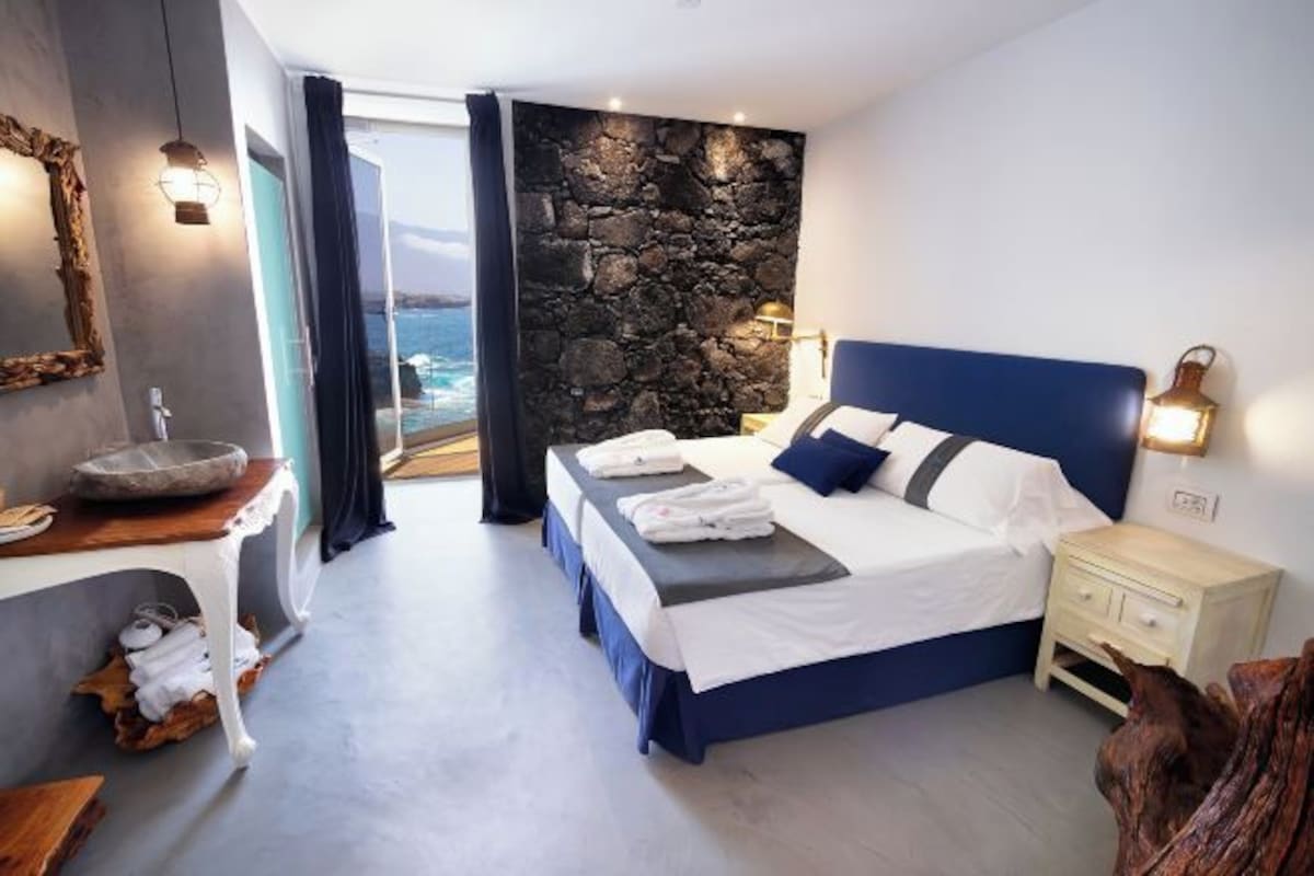 Ein modernes Schlafzimmer mit Steinwand, einem großen Bett mit blauer Bettwäsche, einem Nachttisch und Blick auf das Meer.