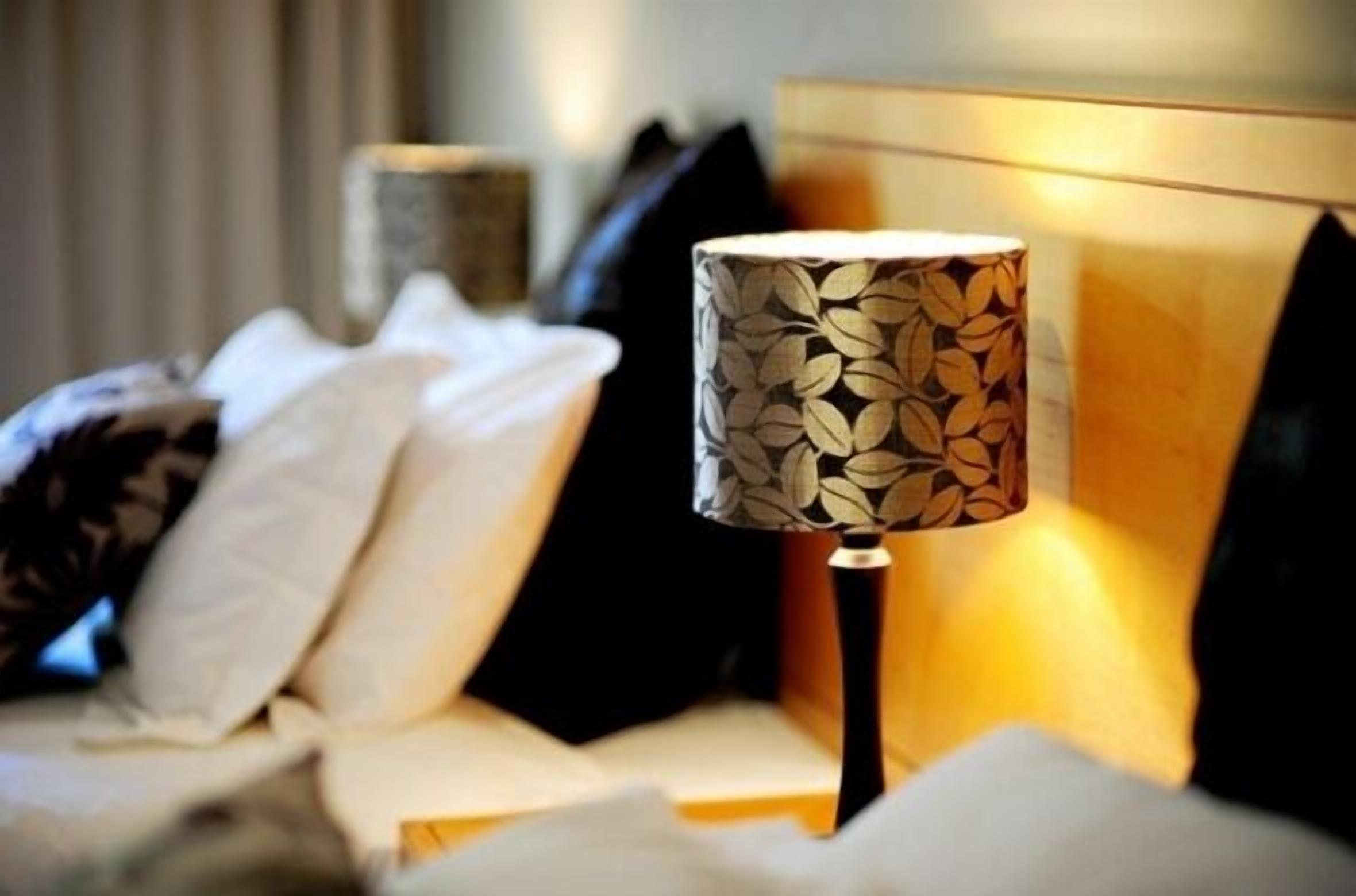 Una cama con ropa de cama blanca y negra, una lámpara con un estampado de hojas y un cabecero de madera.