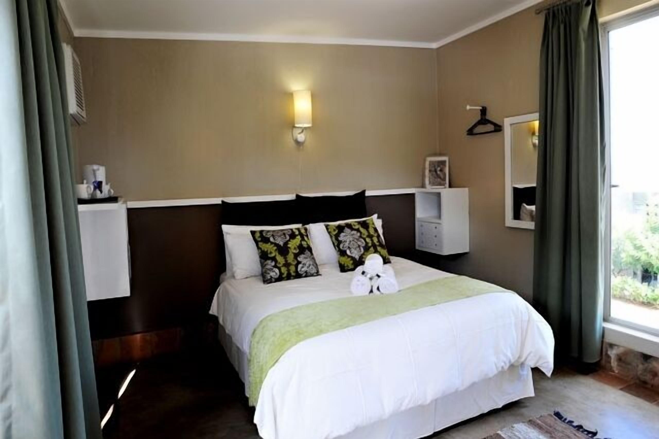 A hotel bedroom with a bed, a nightstand, a wall-mounted lamp, and a window with curtains.