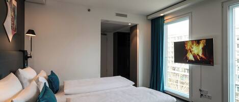 A modern hotel room with a large bed, a flat-screen TV mounted on the wall, and a view of the city through the windows.