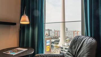 Ein Zimmer mit Ledersessel, einem kleinen Tisch mit einer Zeitschrift, einer Lampe und einem Fenster mit Blick auf einen Turm und Gebäude.