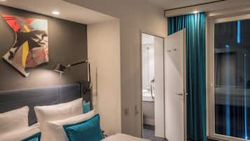 A hotel room with a bed, a desk, a chair, a desk lamp, a painting on the wall, and a bathroom visible through an open door.
