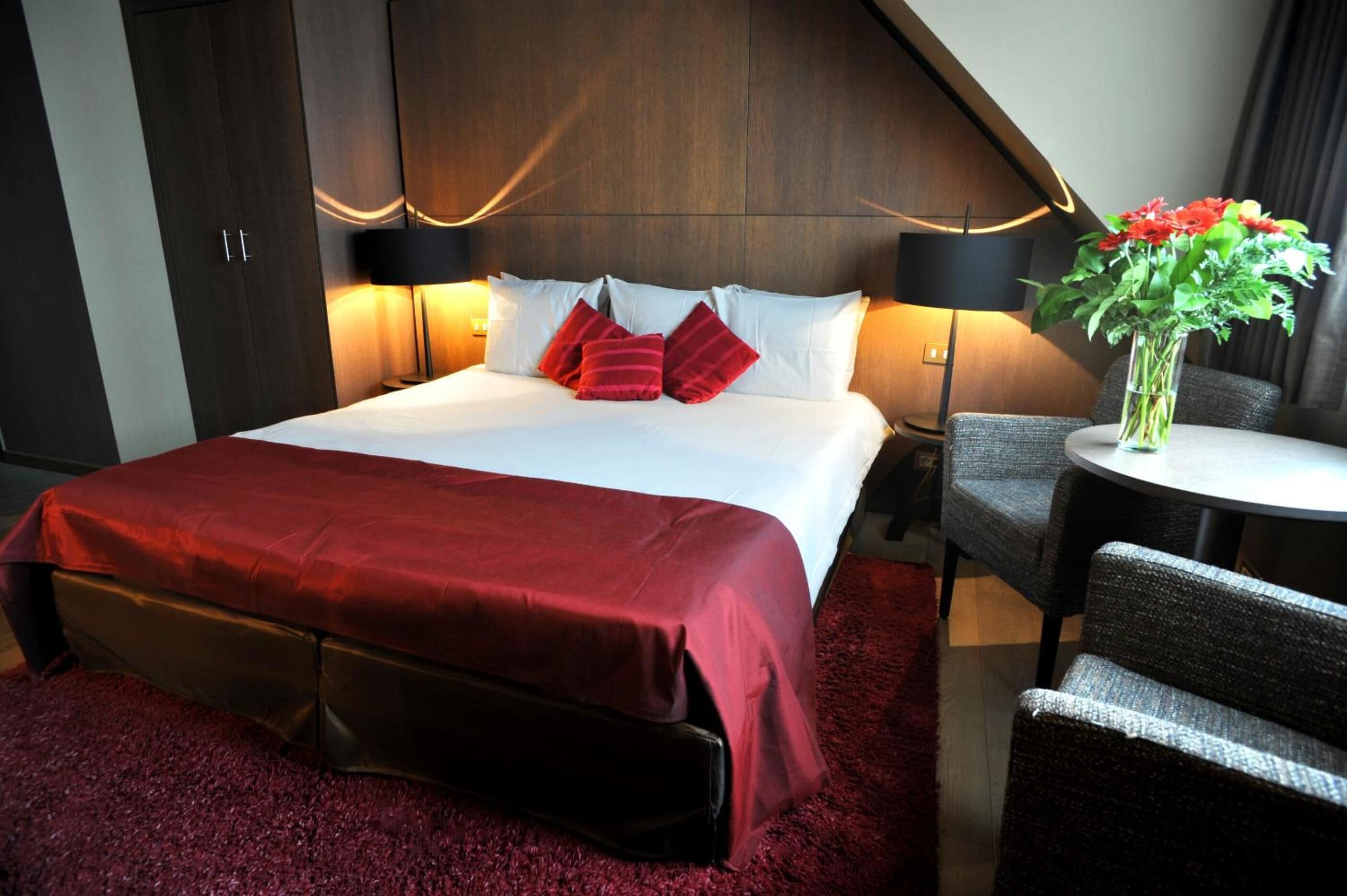 Ein Hotelzimmer mit einem großen Bett, roter Bettwäsche, einem kleinen runden Tisch, einem Sessel und einer Vase mit Blumen.