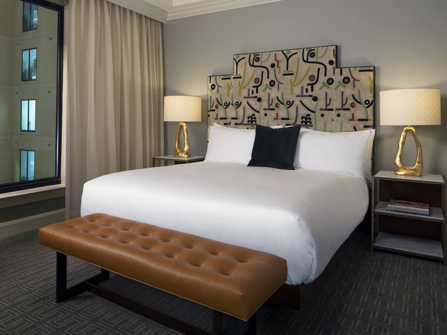 A hotel room with a large bed, two bedside tables with lamps, a bench, and a view of a building through a window.