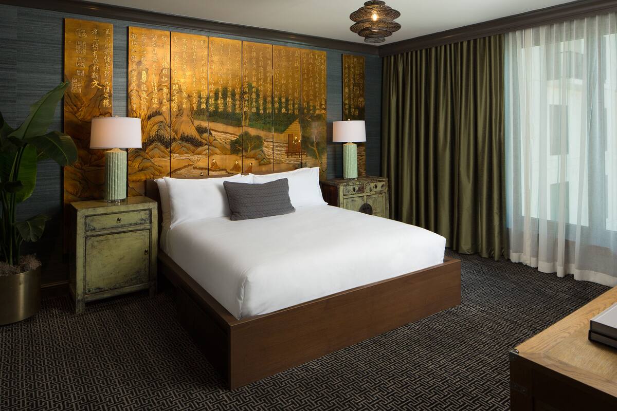 A neatly made bed with a white comforter and a single gray pillow, flanked by two bedside tables with lamps, a large decorative panel with calligraphy, and a view of a building through sheer curtains.
