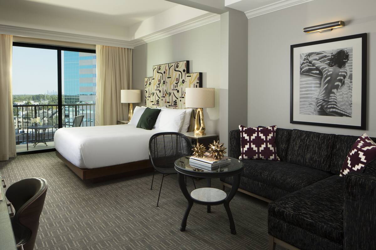 A hotel room with a large bed, a sofa, a small table, and a balcony with a view of the city.