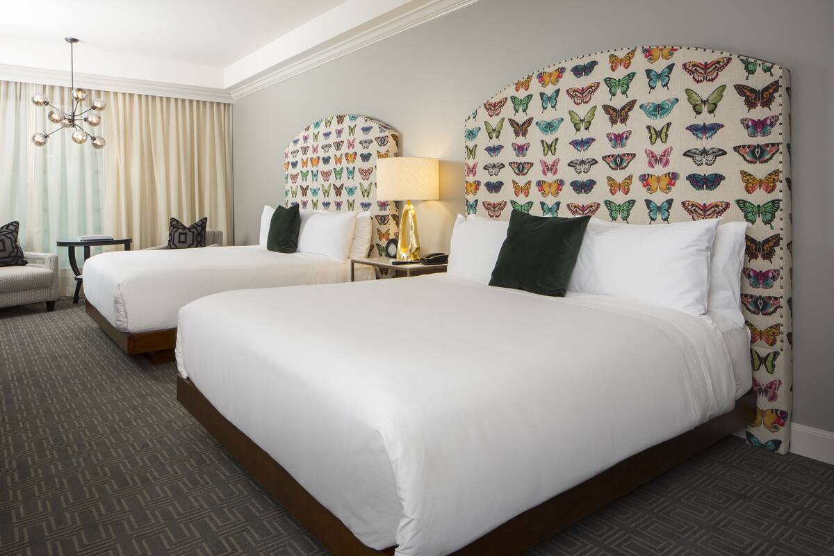 Two double beds with white linens, a chandelier, a small table with a lamp, and a chair.