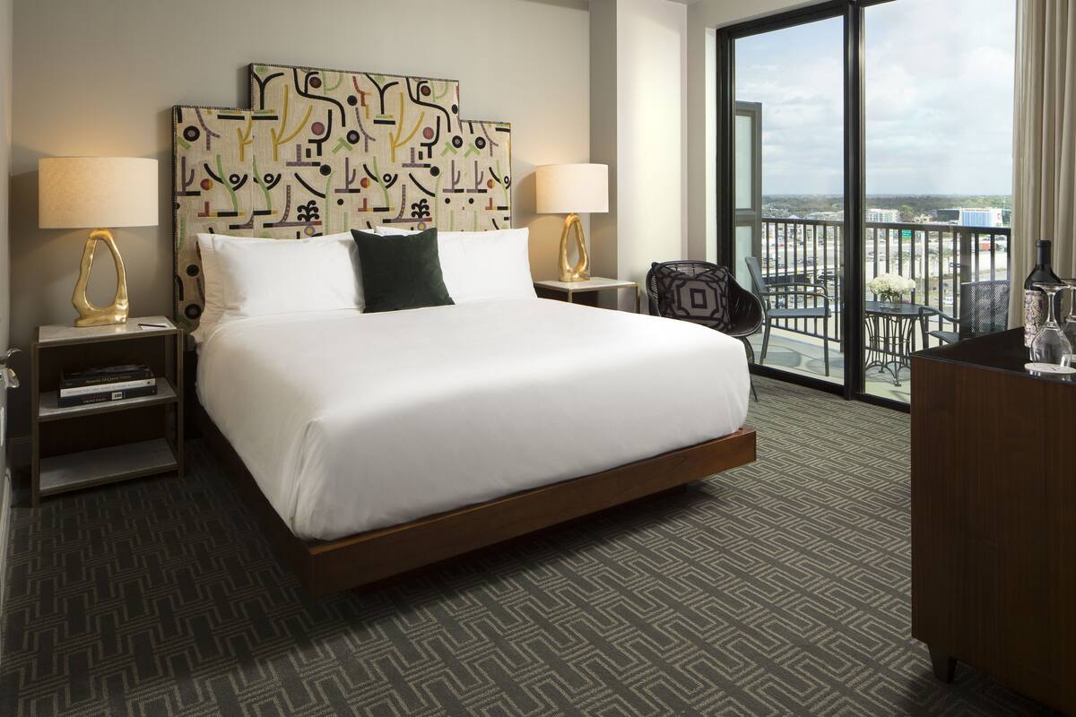 A hotel room with a large bed, two bedside tables with lamps, a chair, a desk, and a view of the city through a large window.