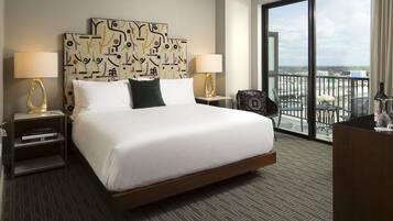 A hotel room with a large bed, two bedside tables with lamps, a chair, a desk, and a view of the city through a large window.