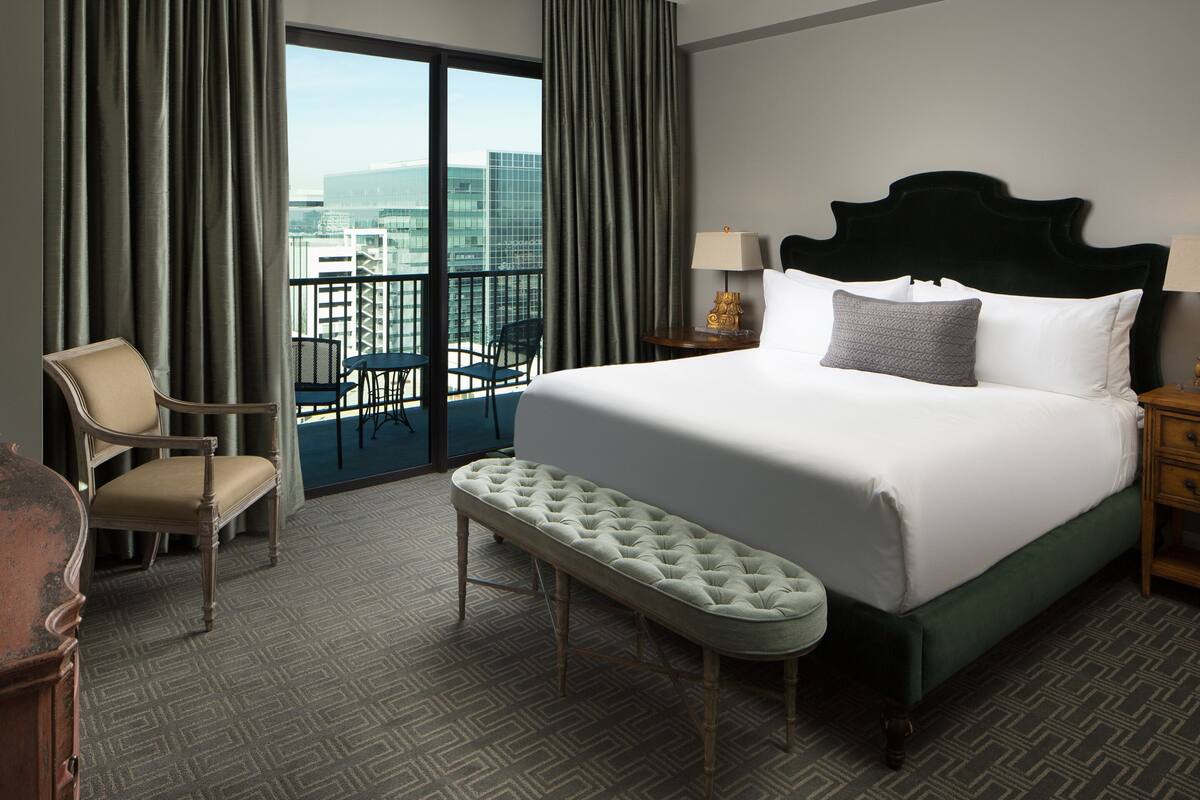 A hotel room with a large bed, a bench, a chair, a nightstand, and a view of the city through a balcony.