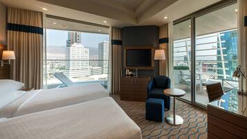 A hotel room with two beds, a TV, a small table, a chair, and a balcony with a view of the city.